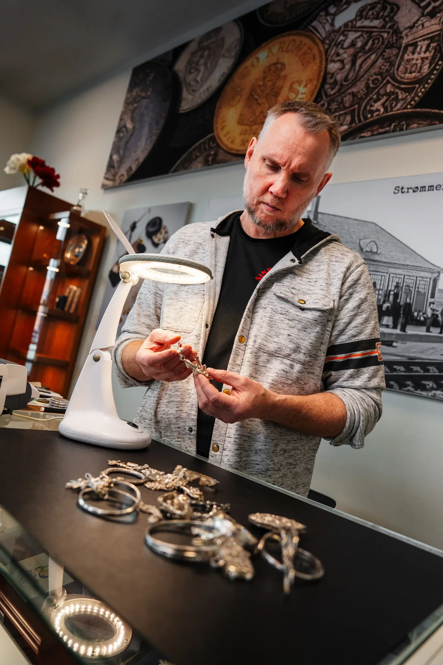 Man meticulously examining a silver chain or bracelet under a magnifying lamp in a shop.