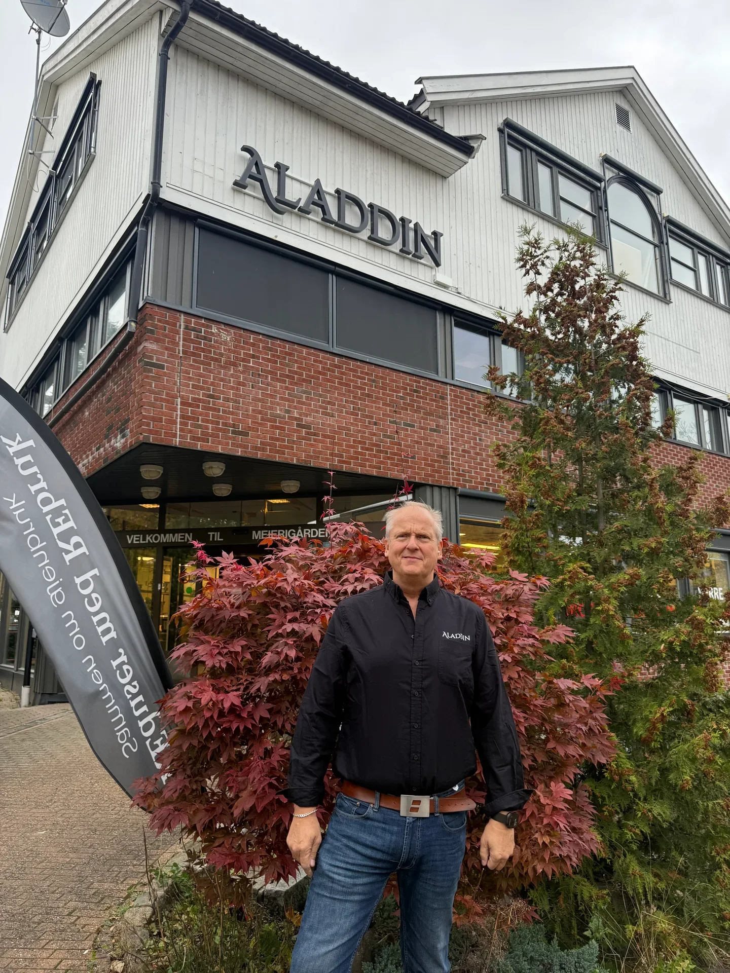 A man in a black shirt and jeans stands before the "ALADDIN" building with red foliage.