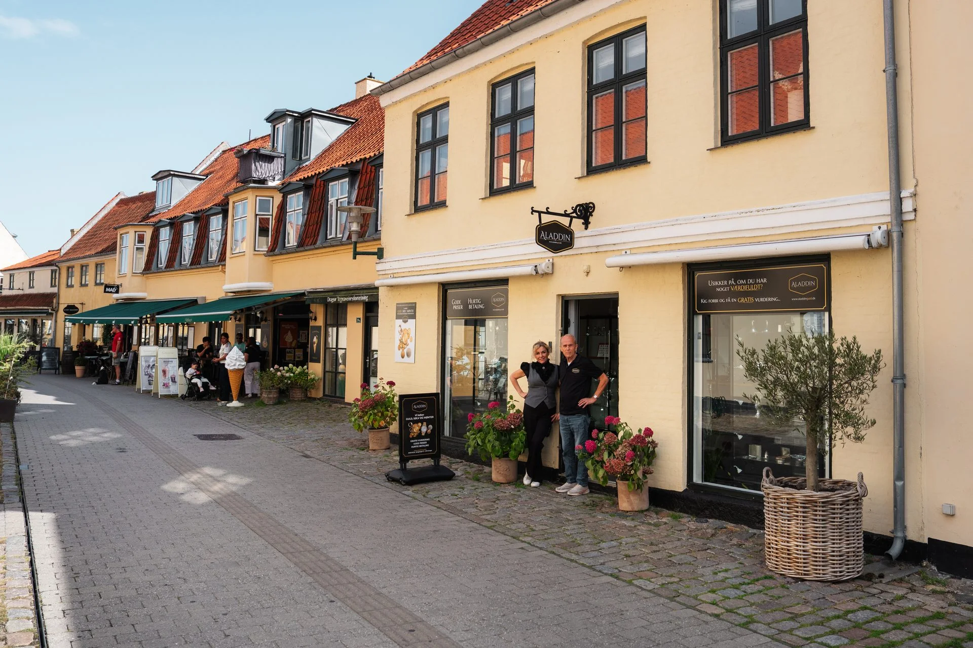 Two people stand outside a yellow shop named "Aladdin" on a charming European street with other shops and buildings.