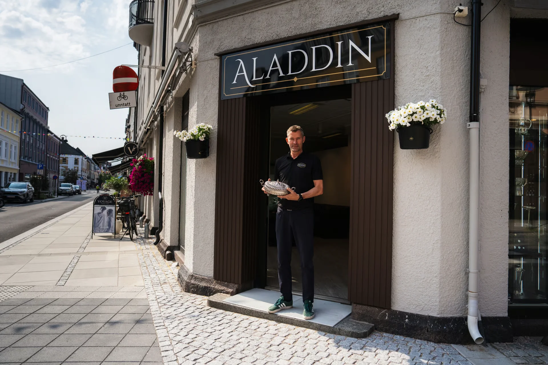 Man holding a silver dish in front of an "ALADDIN" store entrance on a European street.