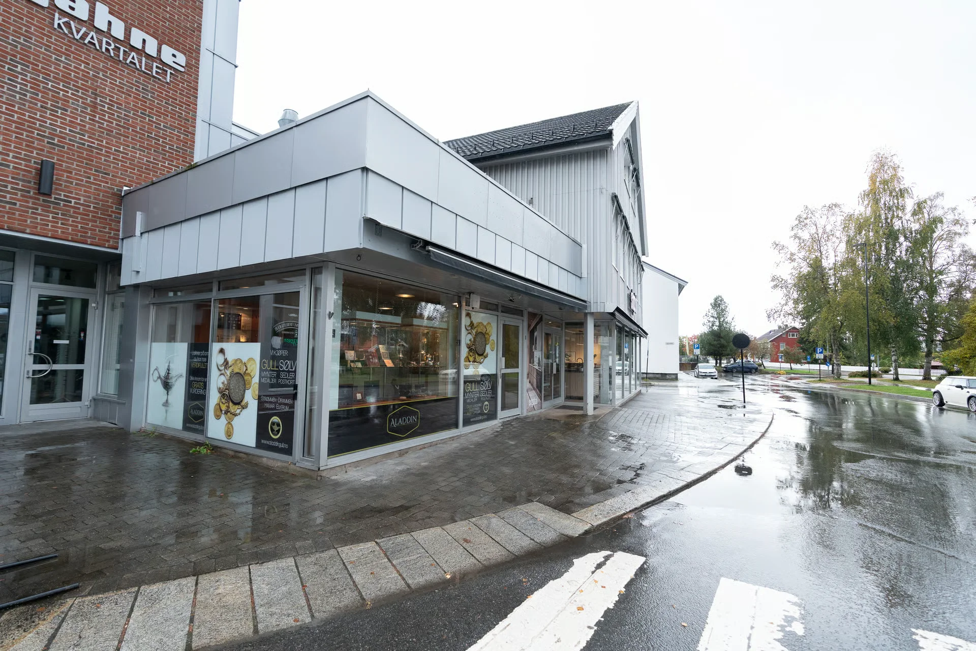 A modern building with an 'Aladdin' storefront on a wet street, featuring brick and light gray siding under an overcast sky.