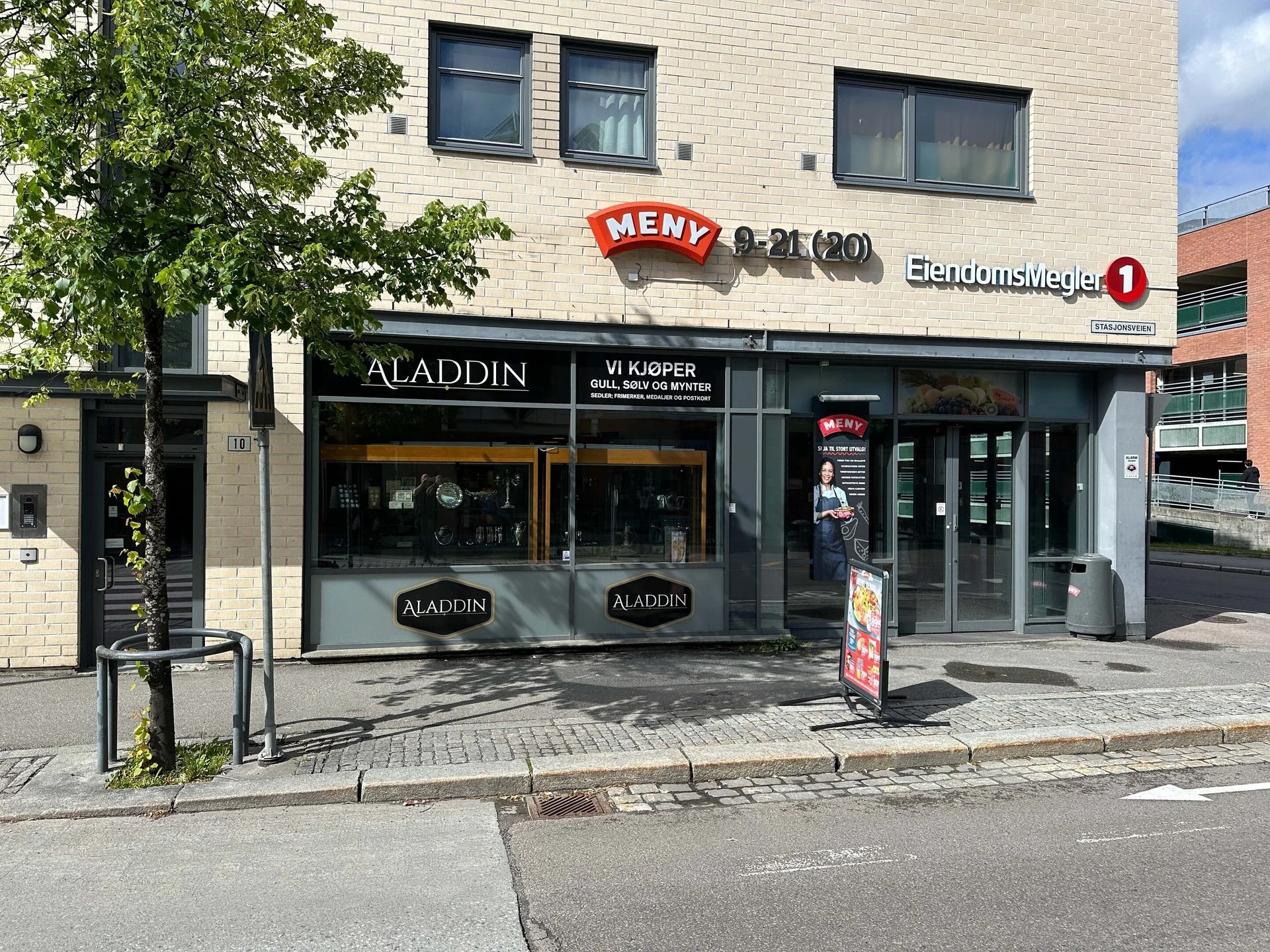 Street view of commercial buildings with a MENY grocery store, Aladdin jewelry, and EiendomsMegler real estate.