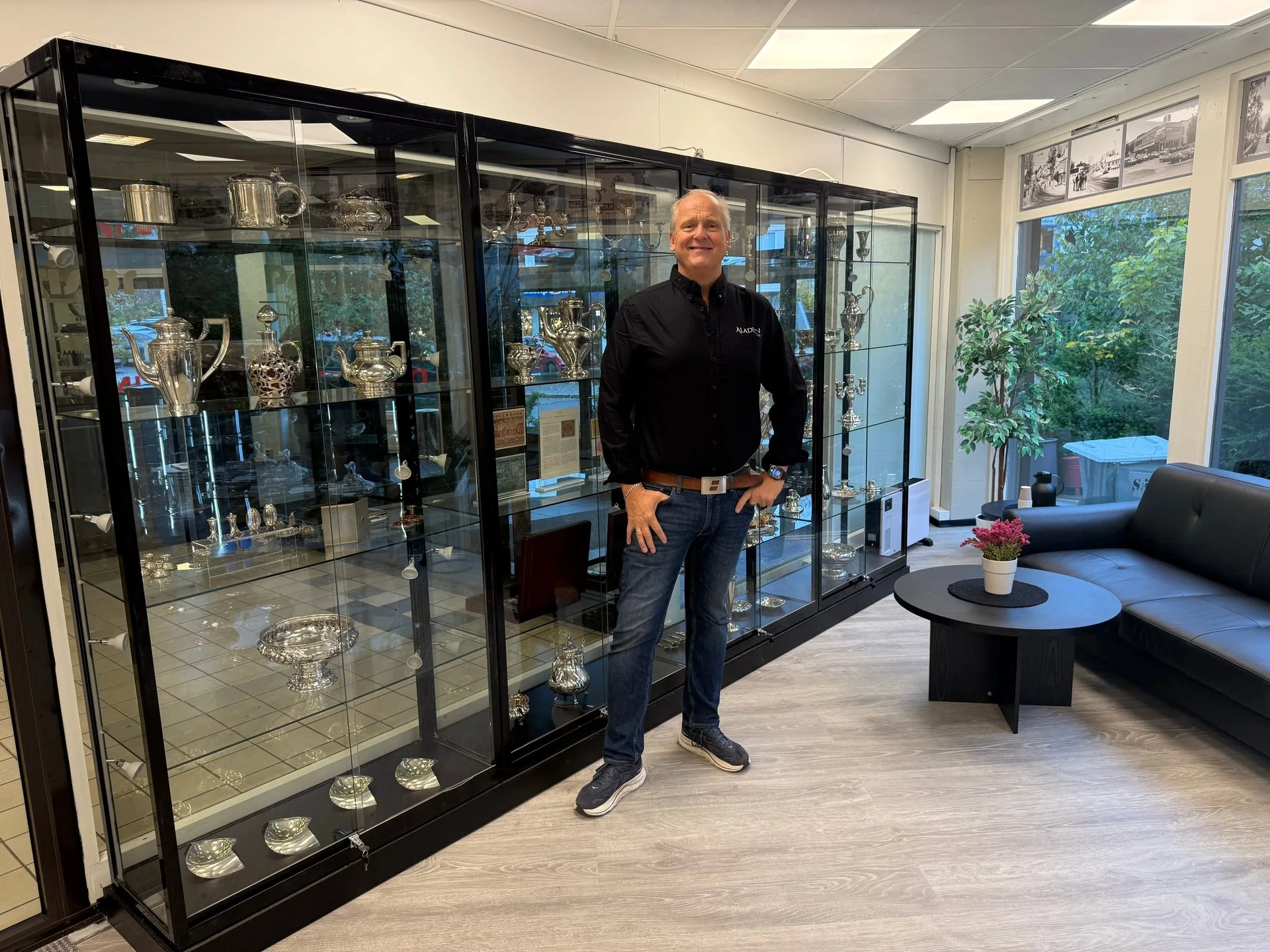 Man smiling in a showroom with display cases filled with silver items and a customer seating area.