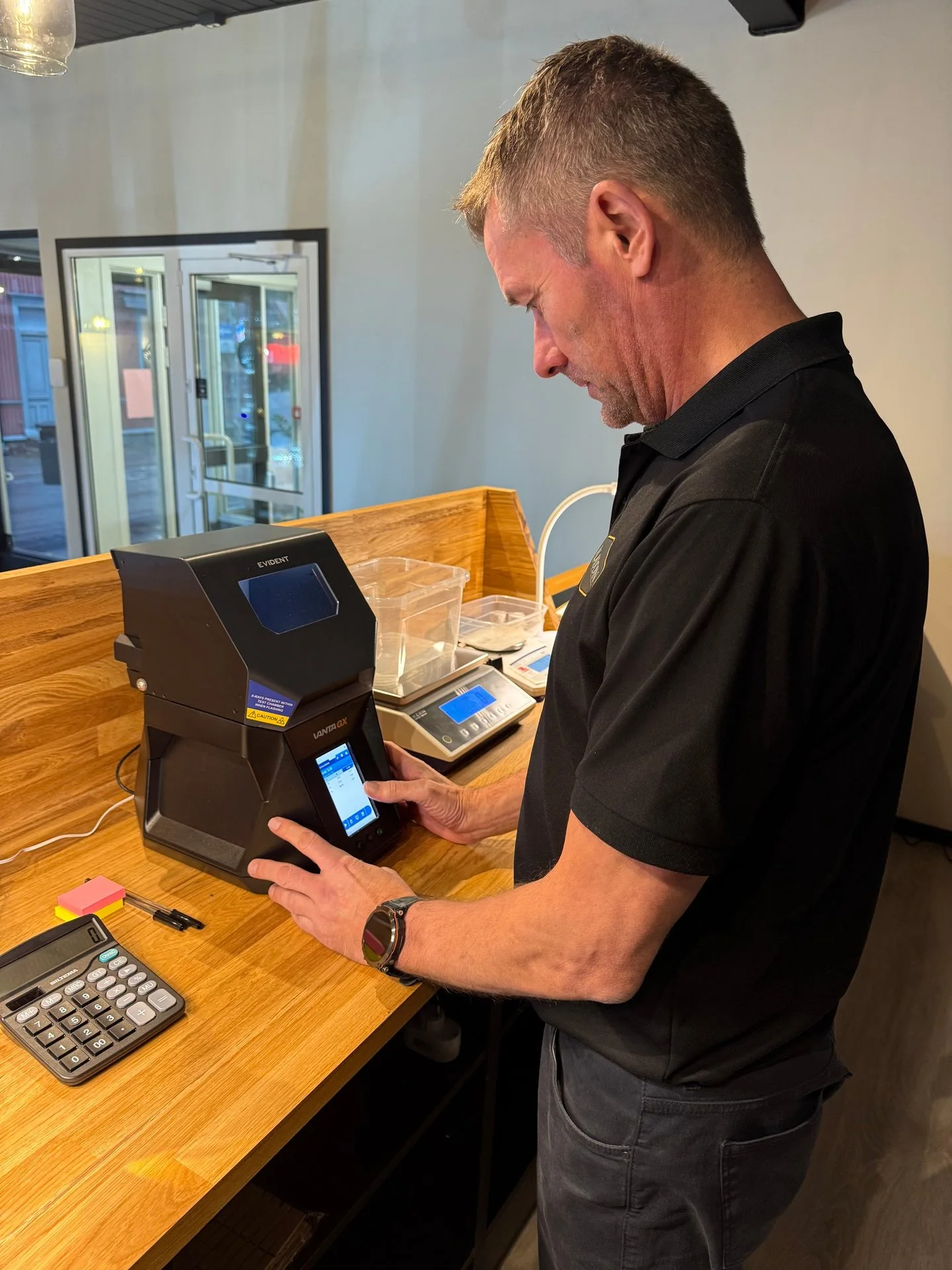Man operates EVIDENT VANTA GX machine on wooden counter with calculator and scales.